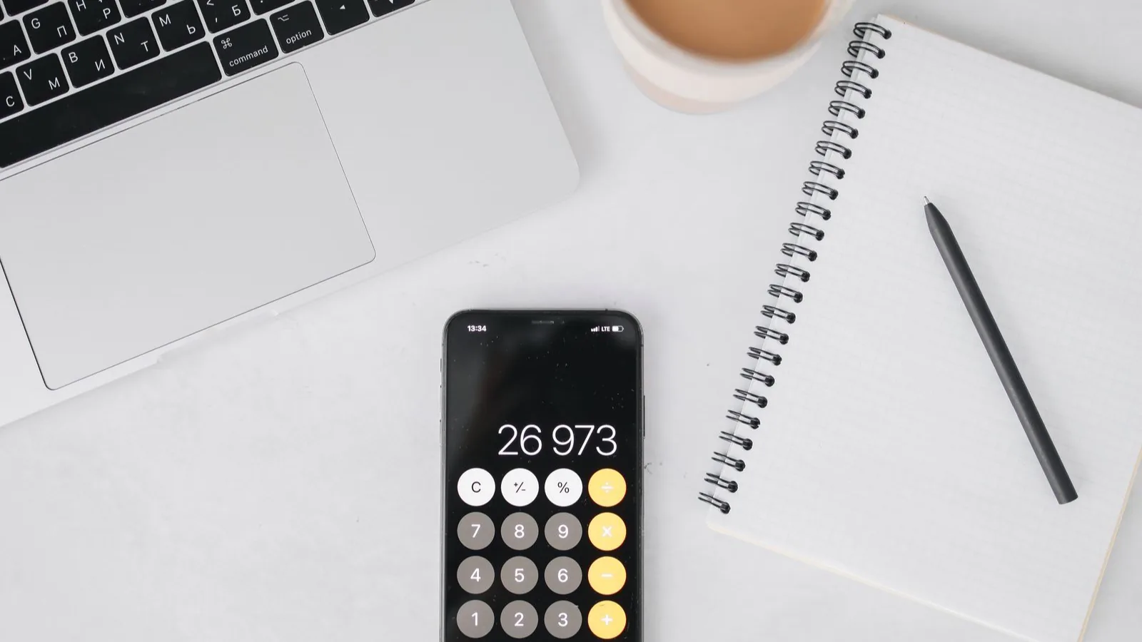 A laptop, a phone showing a calculator and a notebook on a bright white desk